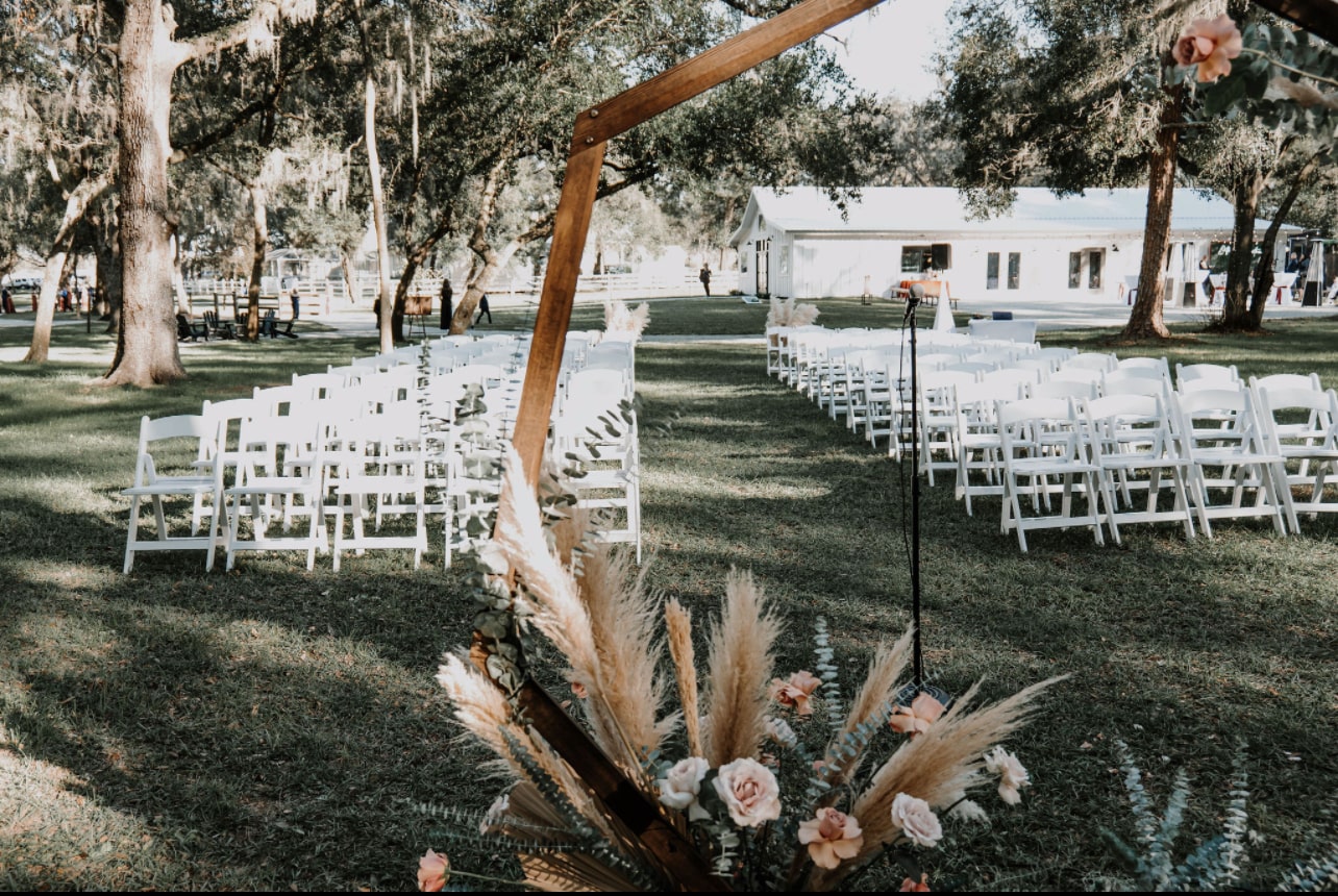 White garden chairs lined up for an outdoor ceremony under the oaks at Lithia Acres, pampas grass on the arch