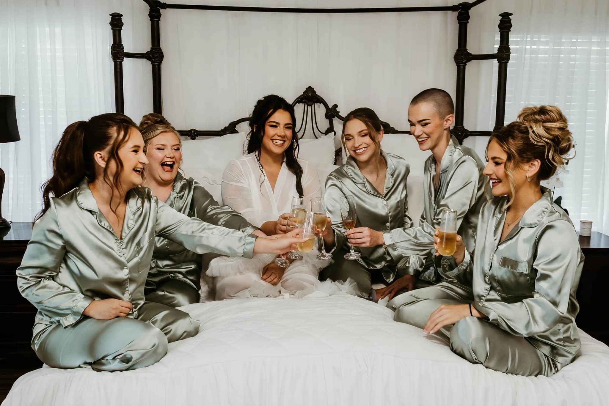 A bride and her bridesmaids in sage satin pajamas toasting on the bed in the Bridal Suite