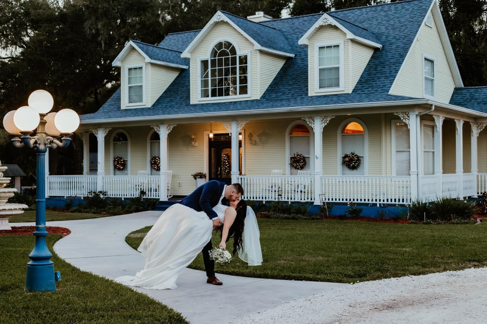 A couple in a dip on the front walk of the white farmhouse with its blue roof and porch